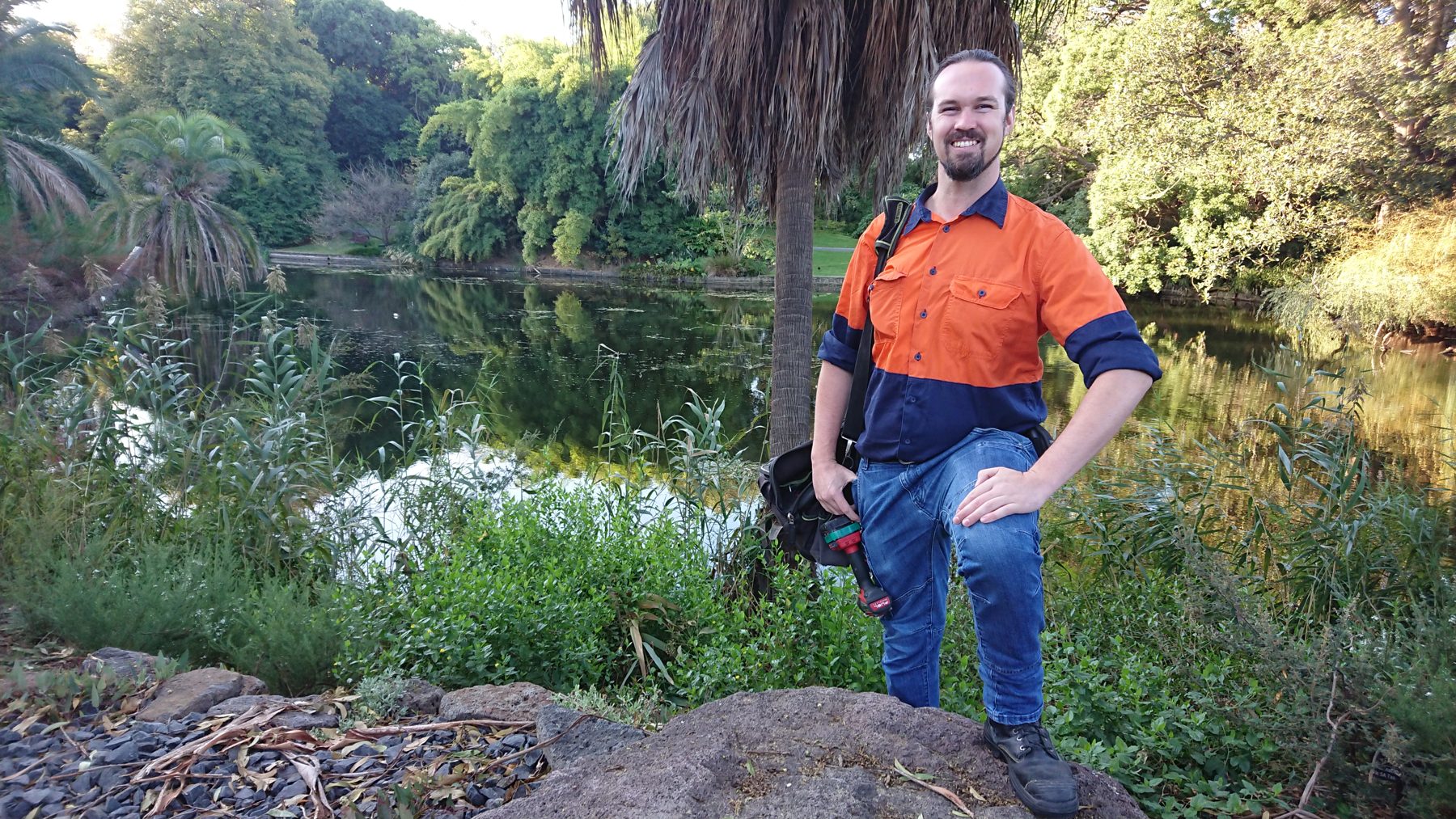 Max Dicks in hi-vis workwear, smiling beside a lake in the Royal Botanic Gardens